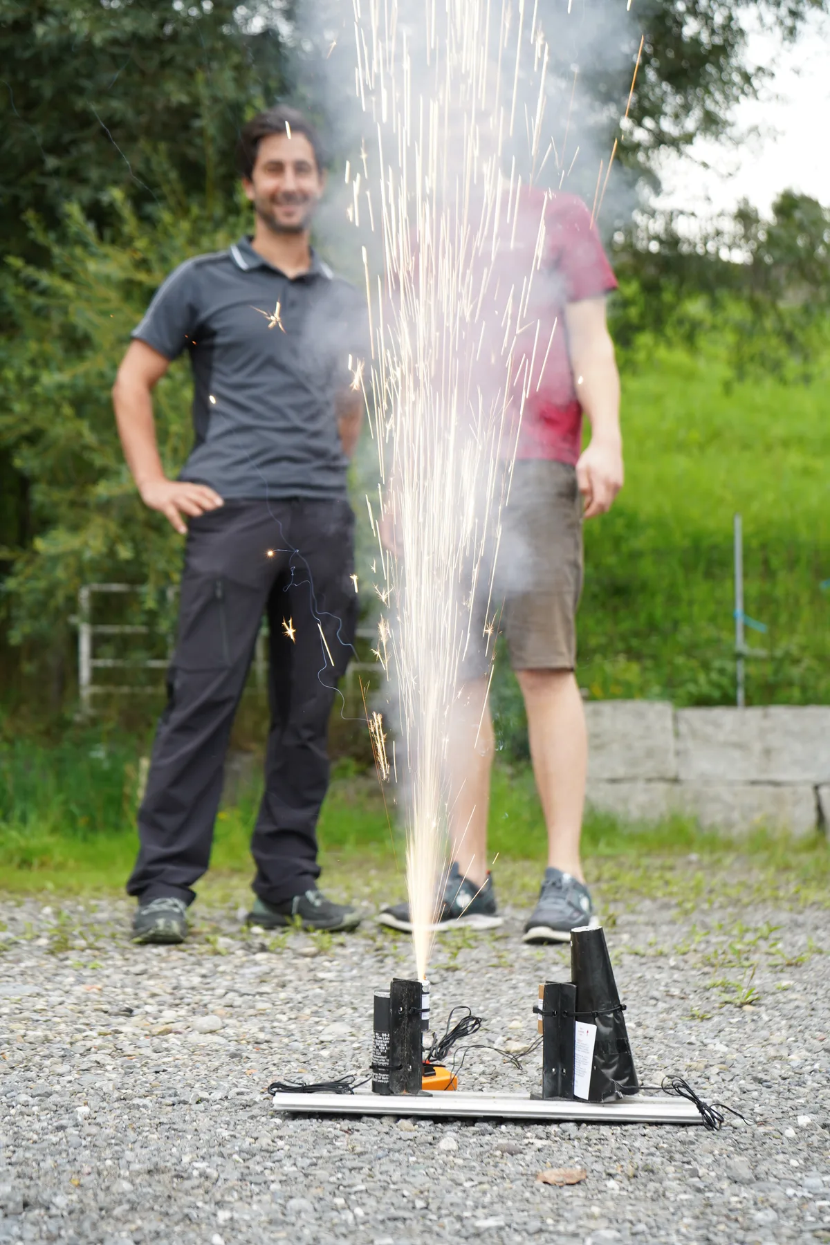 Im Vordergrund wird ein Pyro-Effekt gezündet, hinten stehen zwei Männer und schauen zu.